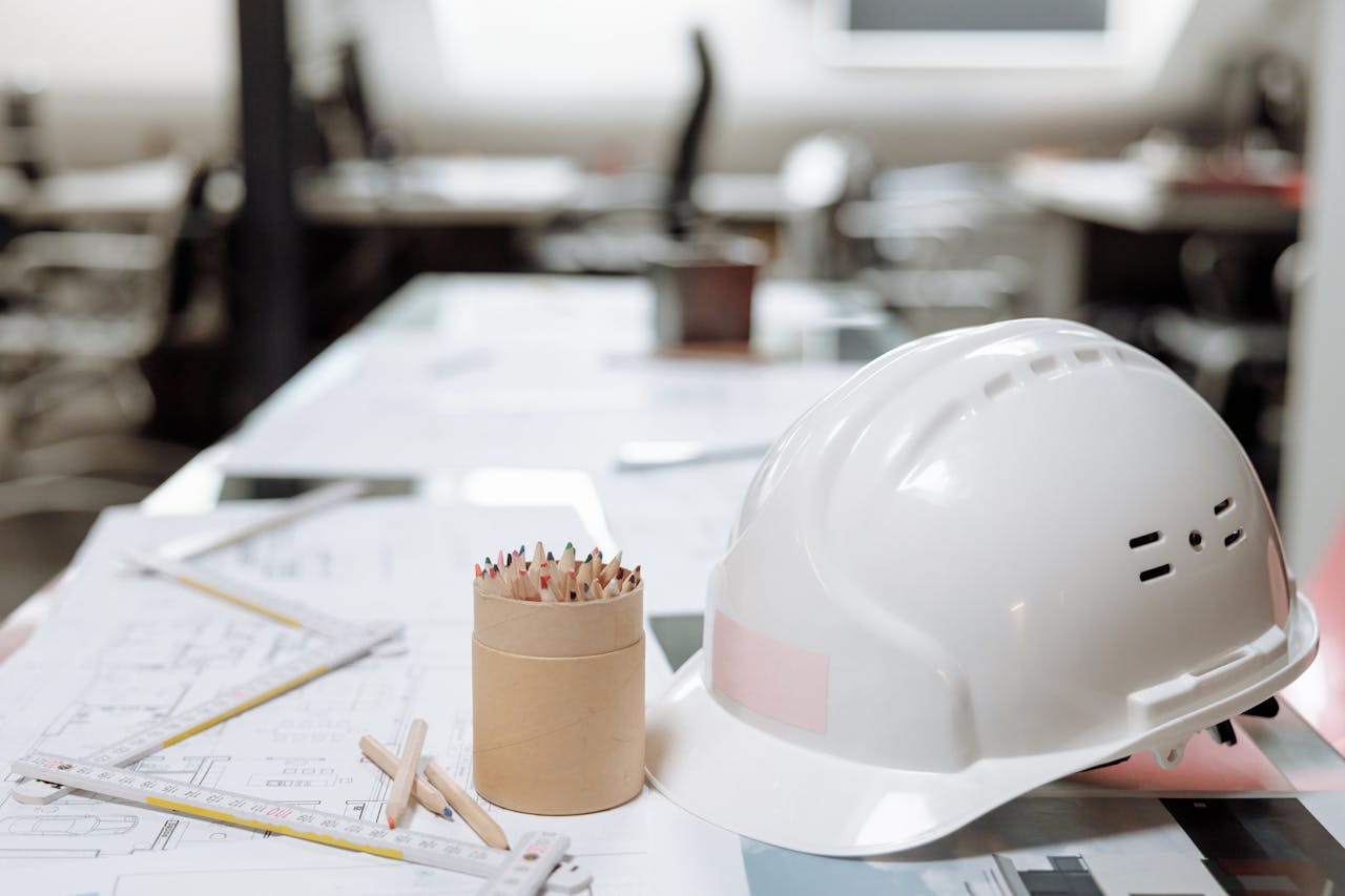 who-we-are Close-up of an office desk with a white helmet, colored pencils, and architectural plans.